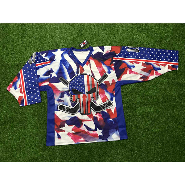 A hockey jersey laid flat on green artificial turf. The jersey has a patriotic red, white, and blue camouflage pattern with a central skull graphic featuring crossed hockey sticks and an American flag motif.