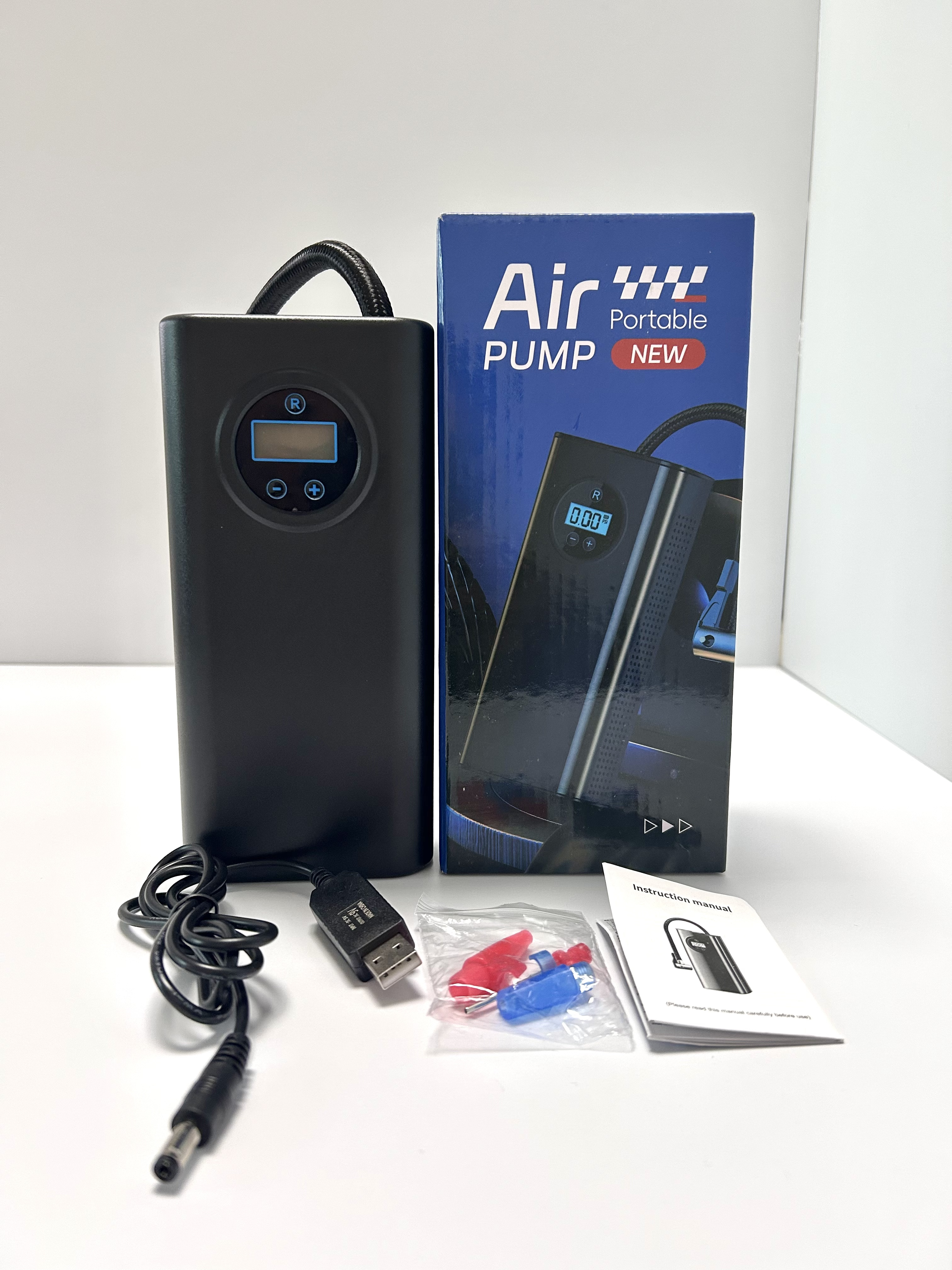 A portable air pump kit displayed on a white surface, including the black pump with digital display, its blue retail box, USB cable, air filter, red and blue air valves, and an instruction manual.