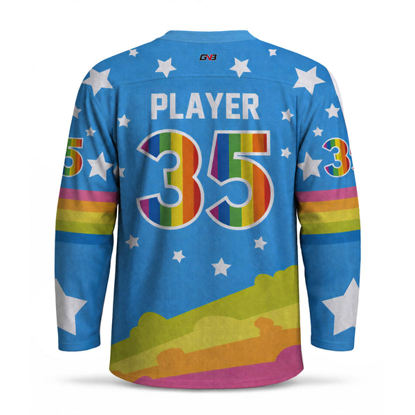 A long-sleeve hockey-style jersey shown from the back on a white background. The jersey is bright blue with white stars, personalized name  and large number in rainbow stripes, and rainbow stripes plus a rainbow mountain graphic near the hem.