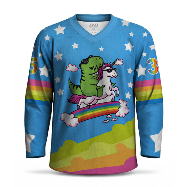 A long-sleeve V-neck hockey-style jersey in bright blue with a whimsical dinosaur-and-unicorn graphic on the chest, white stars, rainbow stripes on the sleeves, and a multicolor cloud pattern near the hem, shown on a plain white background.